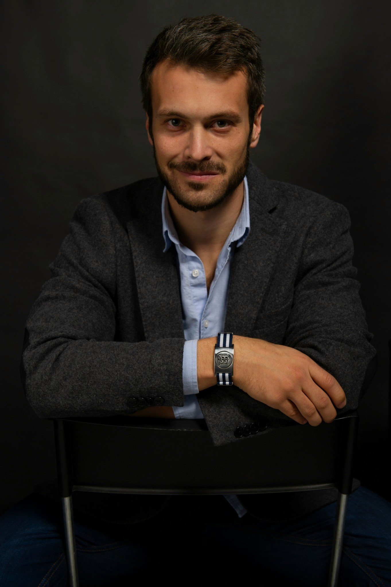 A man with brown hair and a beard sits confidently on a black chair, wearing a grey blazer over a light blue shirt, set against a dark background.
